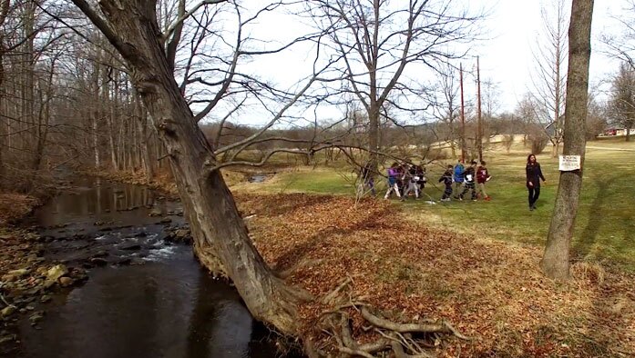 Students are filmed walking in a wooded area near a river as they learn about environmental sciences in an education video production