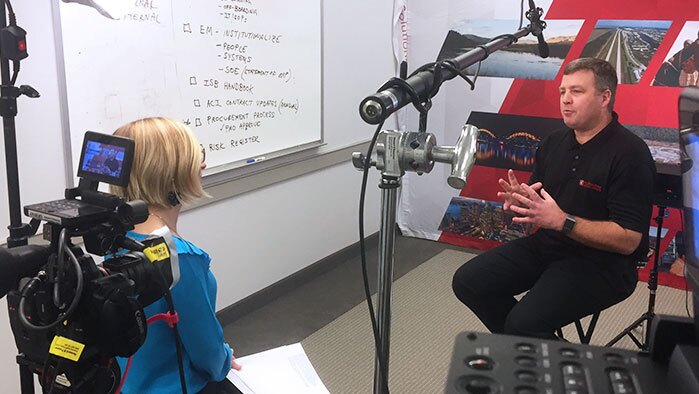 A male employee is interviewd on film by a corporate video production company.