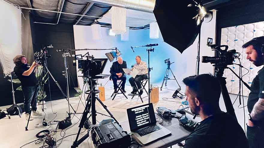 TV hosts are filmed in a New Jersey Production Studio. The studio is filled with cameras, lights, teleprompters and crew.