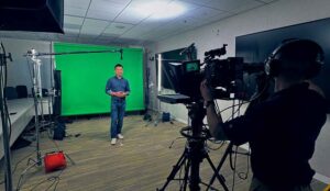 An on camera host in front of a green screen is framed by a camera man with a teleprompter rig for a video production services shoot.